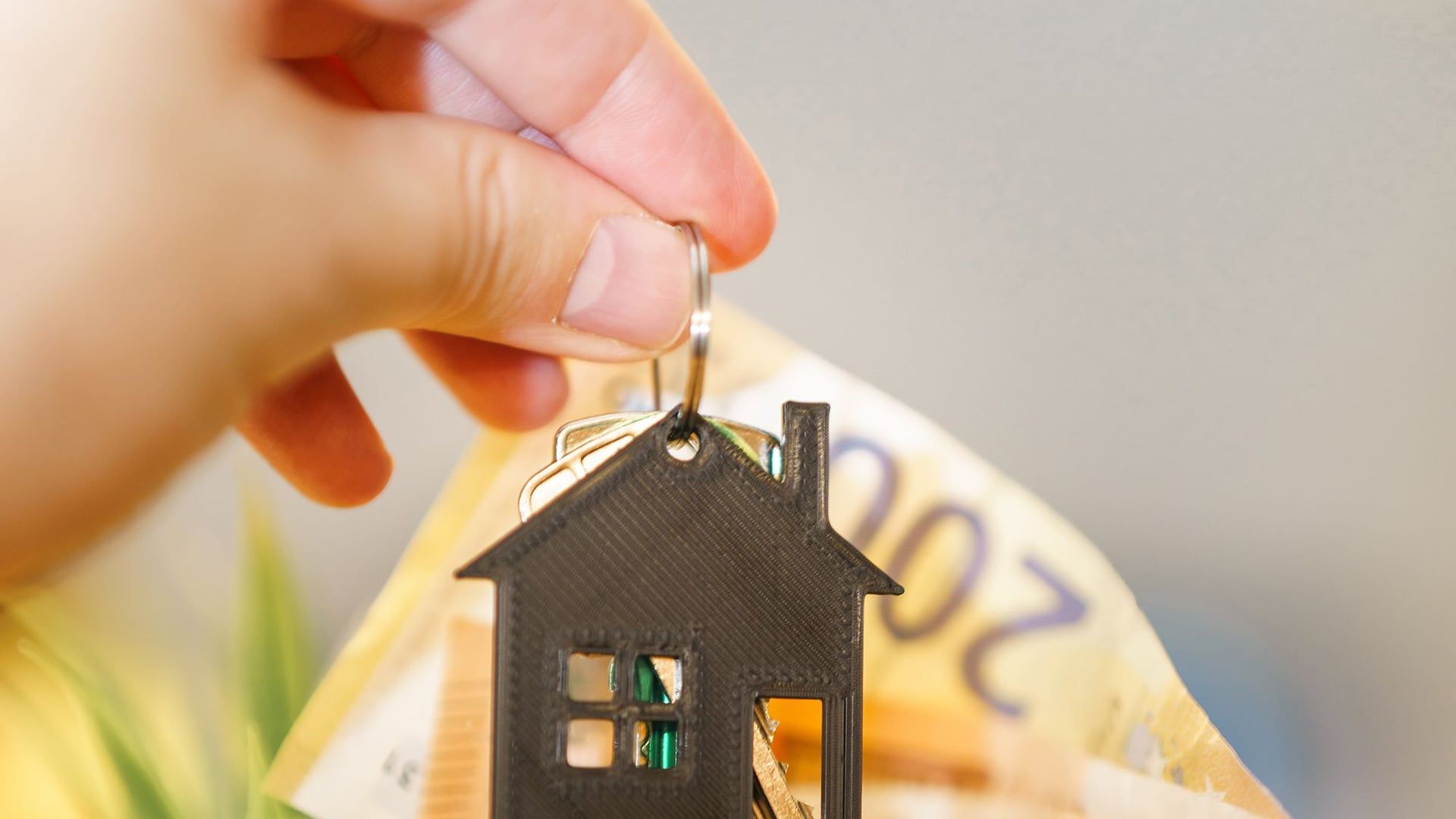 Een hand houdt een 3D-geprinte sleutelhanger in de vorm van een huisje boven een portemonnee met bankbiljetten